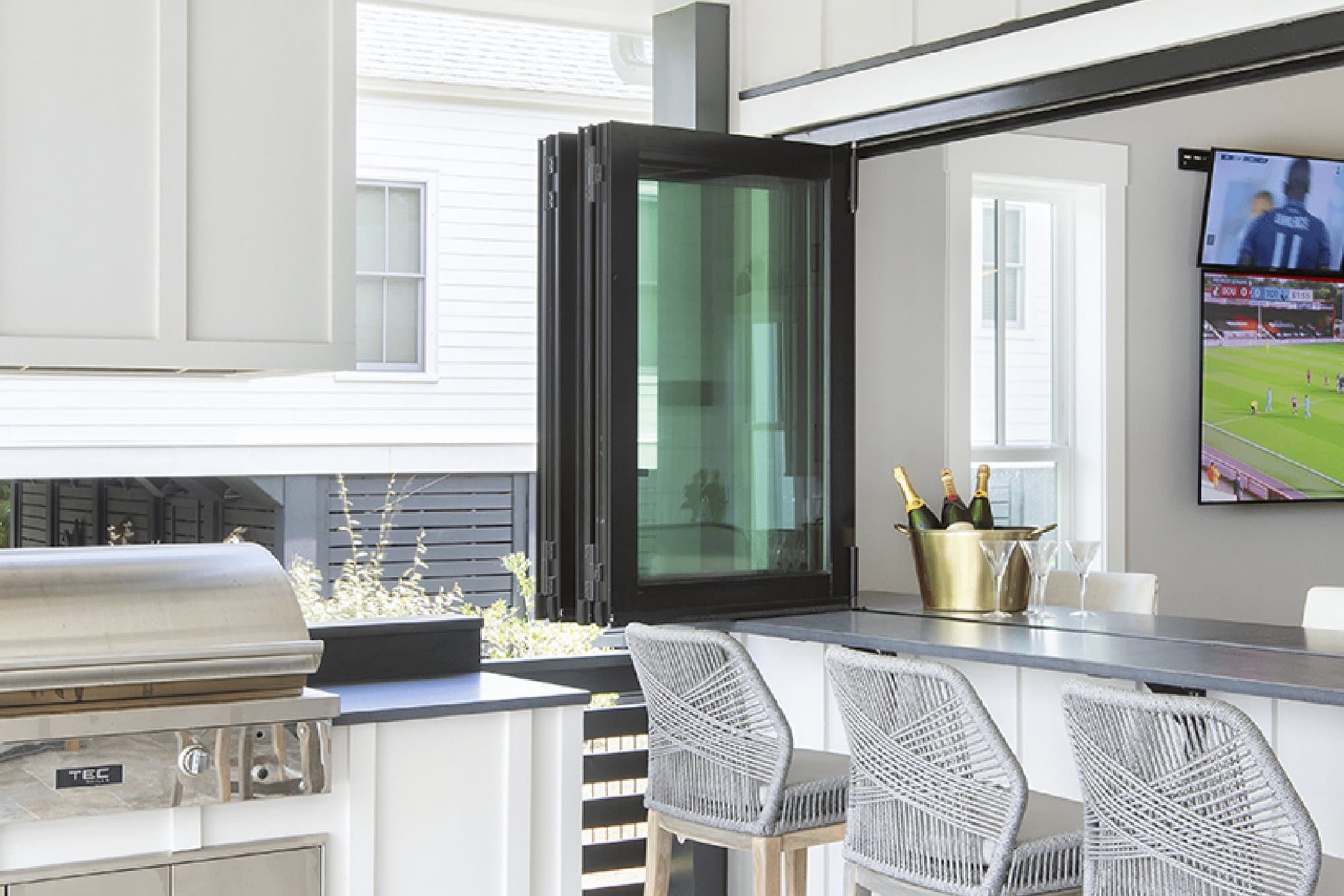 Black-framed Euro-Wall Vista Fold folding glass panels stacked beside an indoor bar counter that opens to an outdoor kitchen and grill