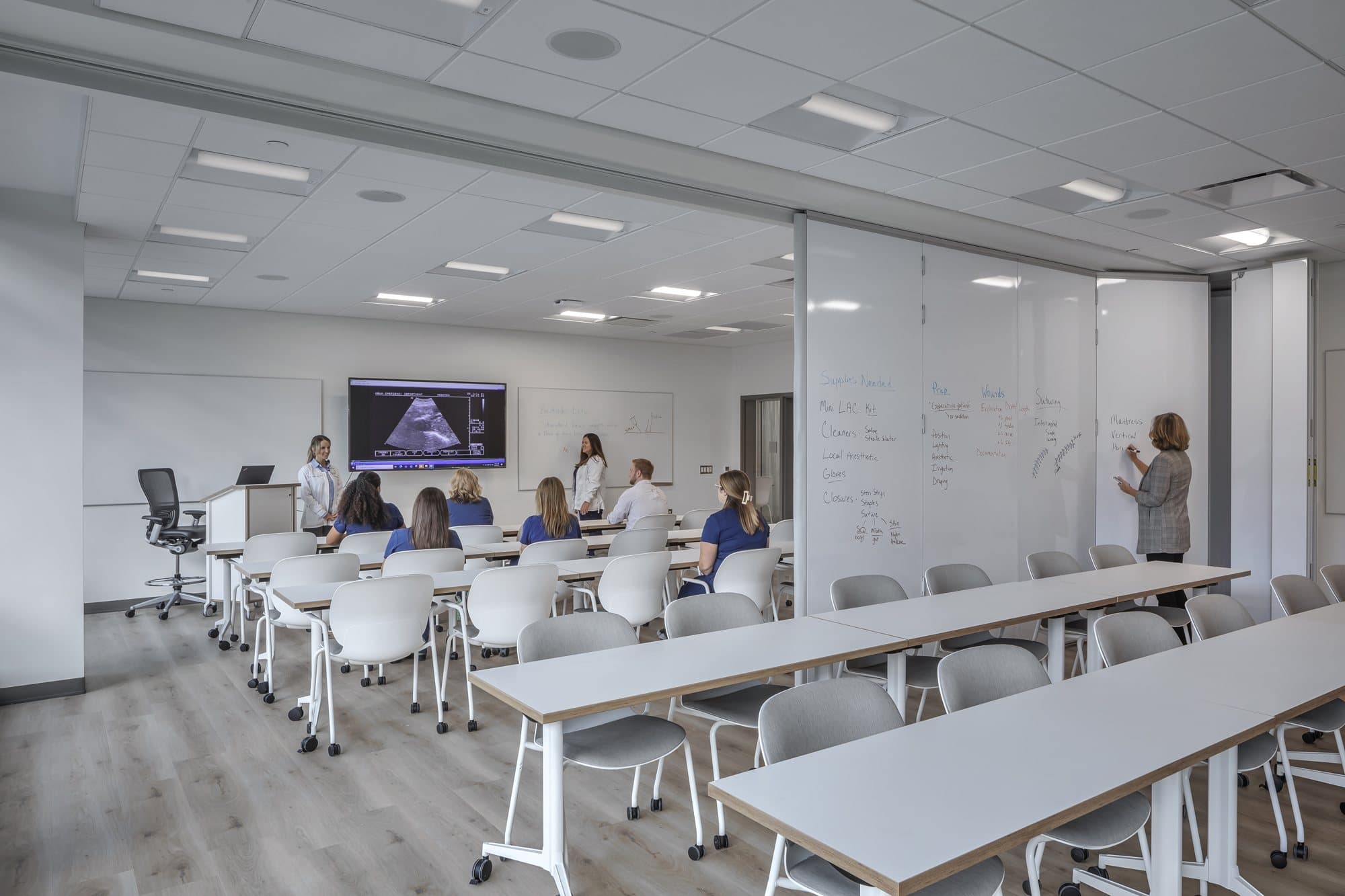 Markerboard-faced Modernfold operable wall panels partly extended across a Corewell Health nurse training classroom while an instructor writes on the panel surface