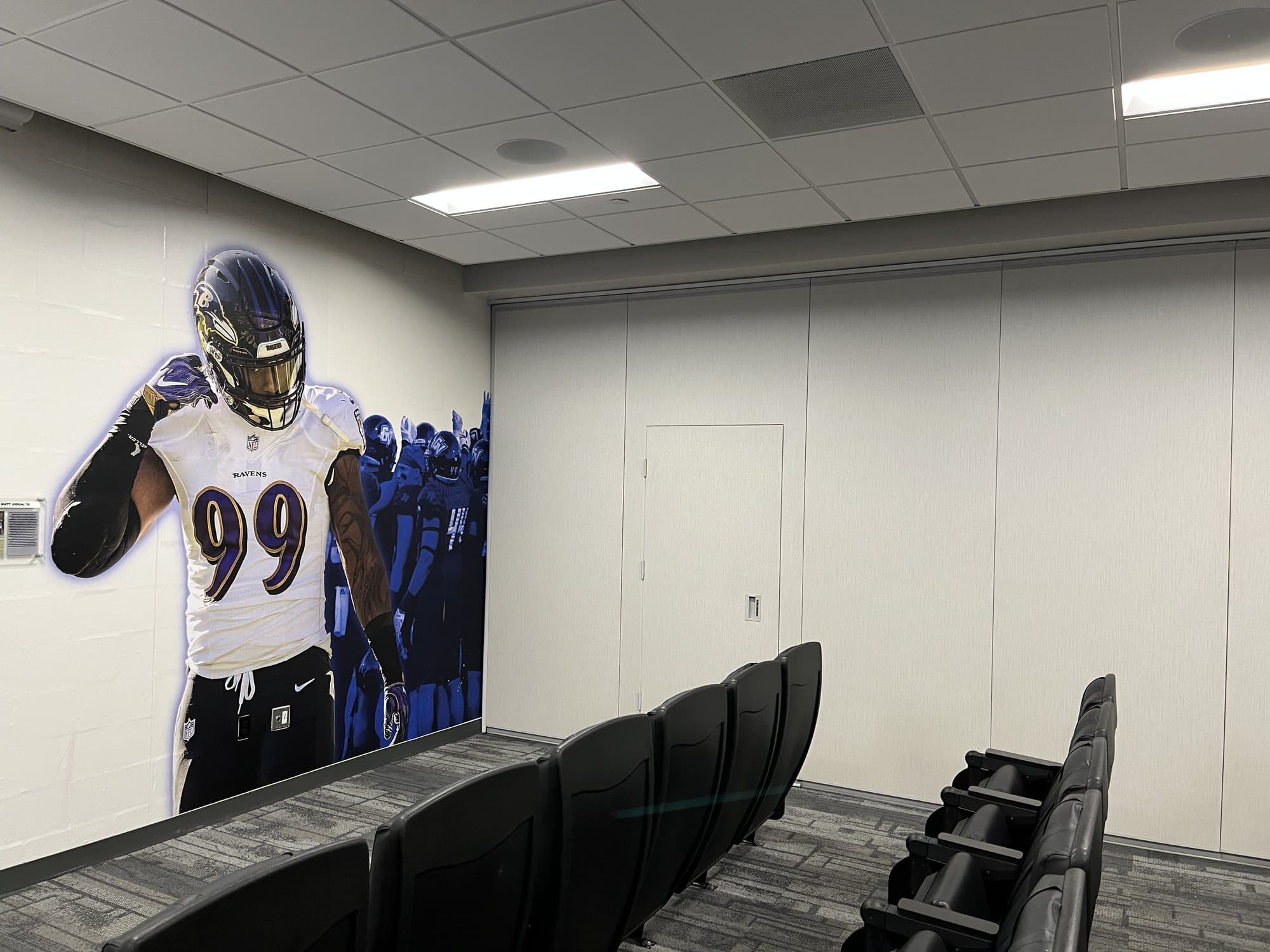 Closed Modernfold operable wall with an in-panel pass door in the GVSU football meeting and film room, beside theater seating and a Ravens mural