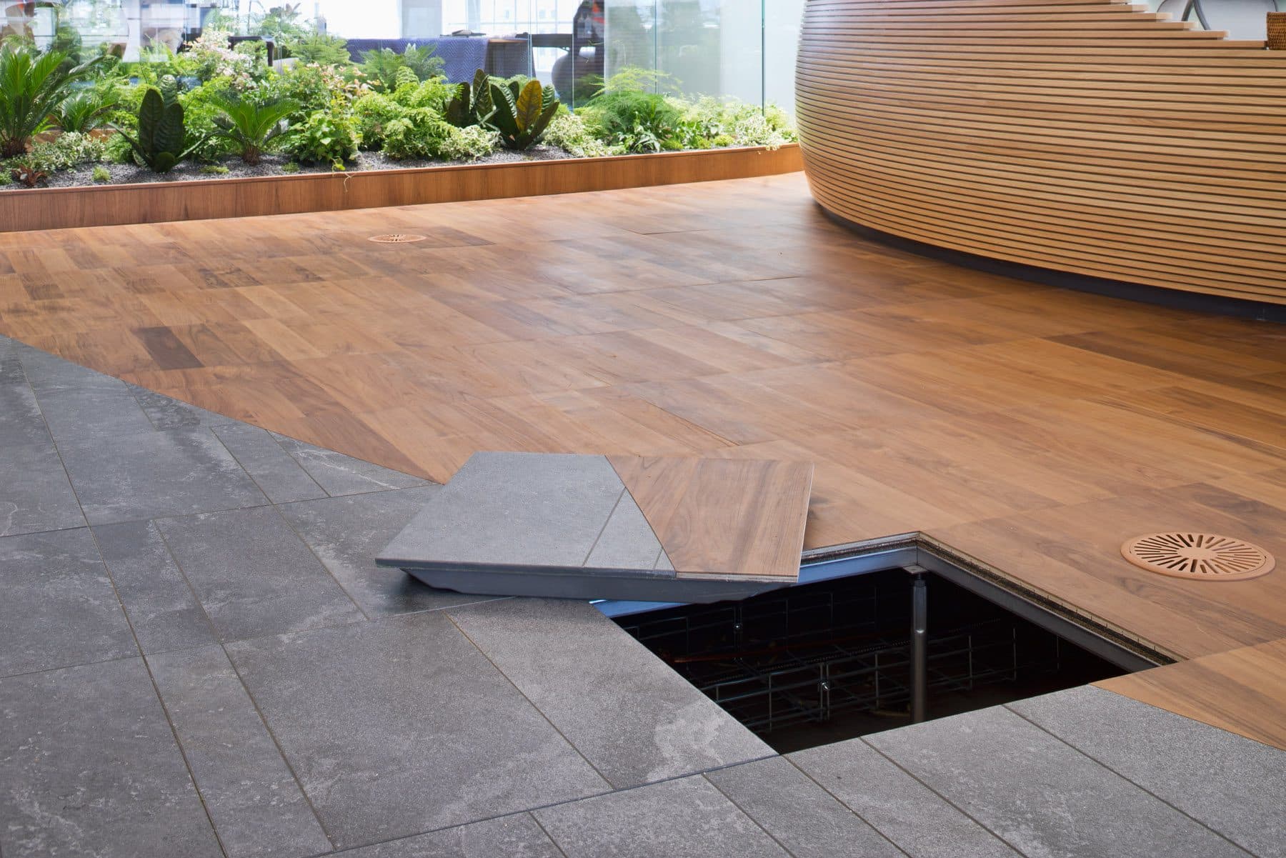 Wood-finish raised access floor in an office with one panel lifted out, showing the open underfloor space