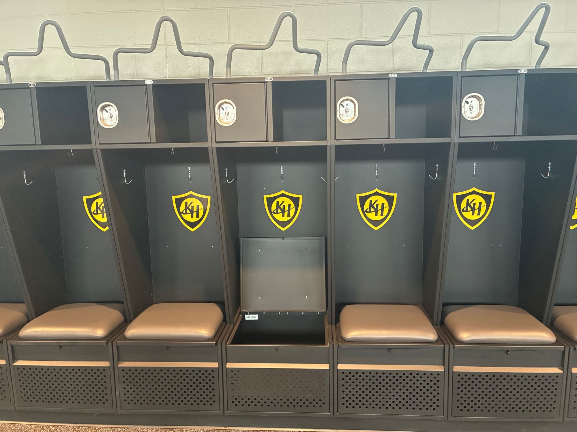 Five charcoal Athletic Edge welded steel athletic lockers with gold KH shield logos, upper cubbies with recessed padlock hasps, coat hooks and padded gray seats, one seat lifted to open the foot locker