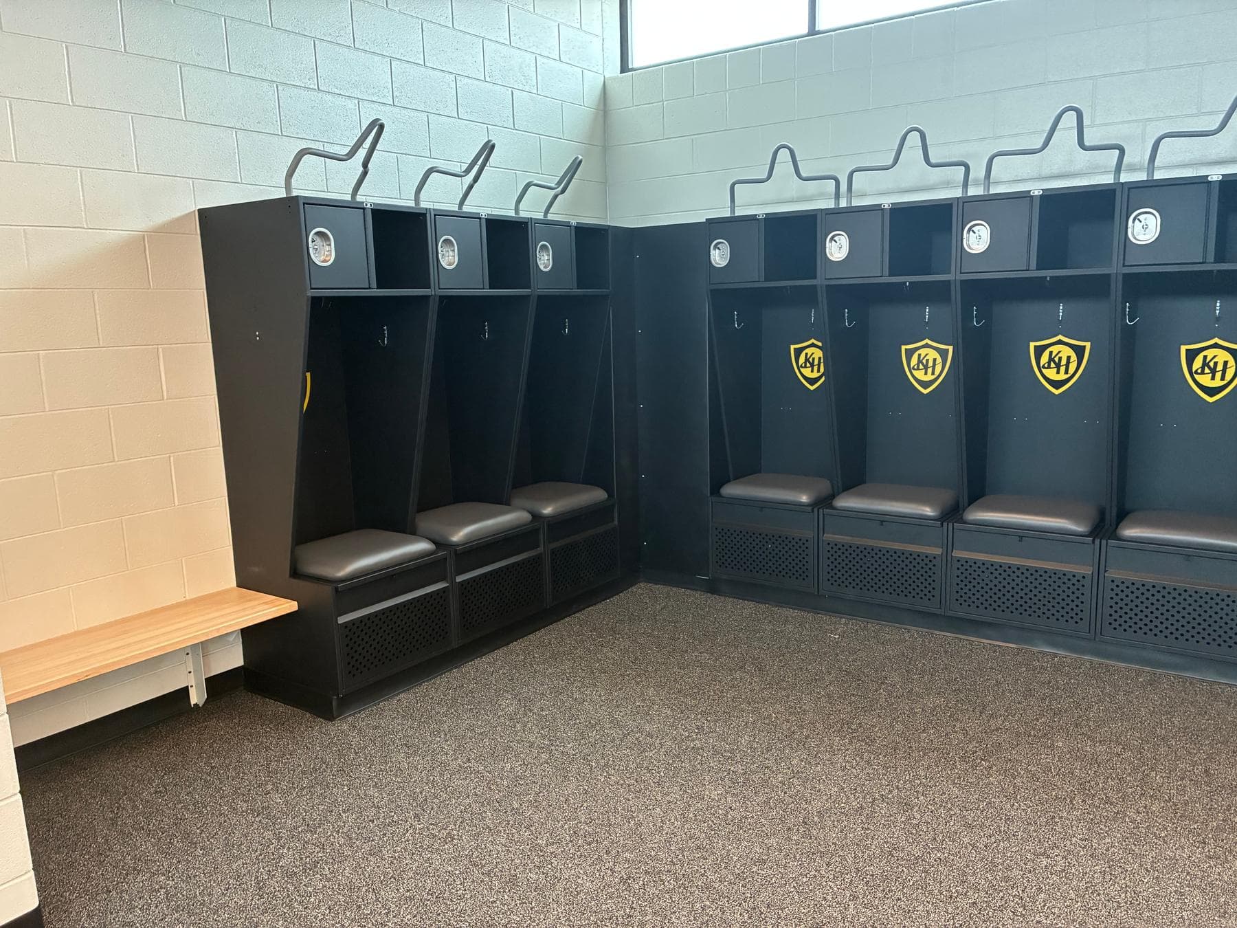 Corner run of charcoal Athletic Edge athletic lockers with gold KH shield logos, padded seats and vented foot-locker fronts, beside a wood bench