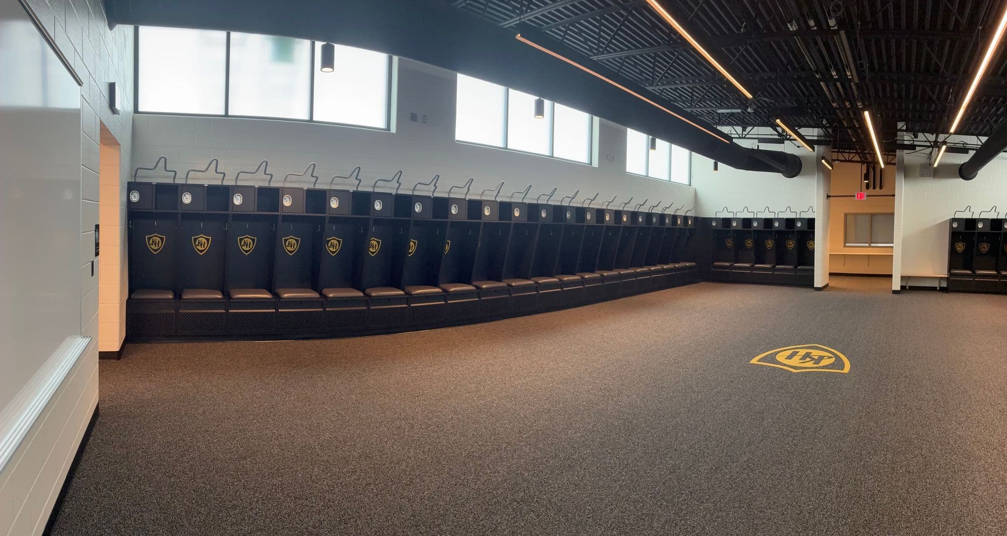 Panorama of the Kenowa Hills High School football locker room, with a long run of charcoal Athletic Edge athletic lockers, each with a gold KH shield logo, and a KH logo inlaid in the floor